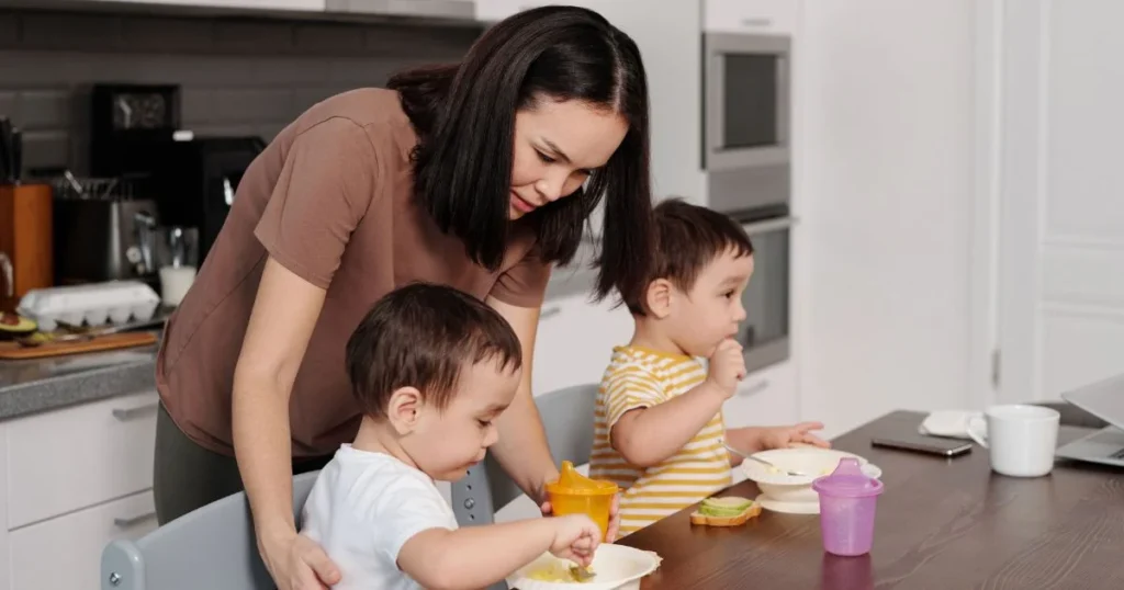 A mother gently assists her young toddler with a sippy cup at the kitchen table while another toddler eats nearby.   